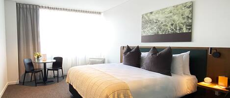 Habitación de hotel con una cama grande, un escritorio con dos sillas, una mesita con un florero y un cuadro en la pared.