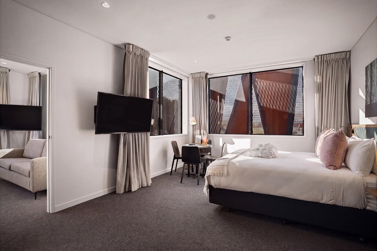 A hotel room with a bed, a desk, a chair, a TV, and large windows.