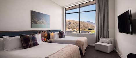 A hotel room with two beds, a flat-screen TV, a large window with a mountain view, and a small table.