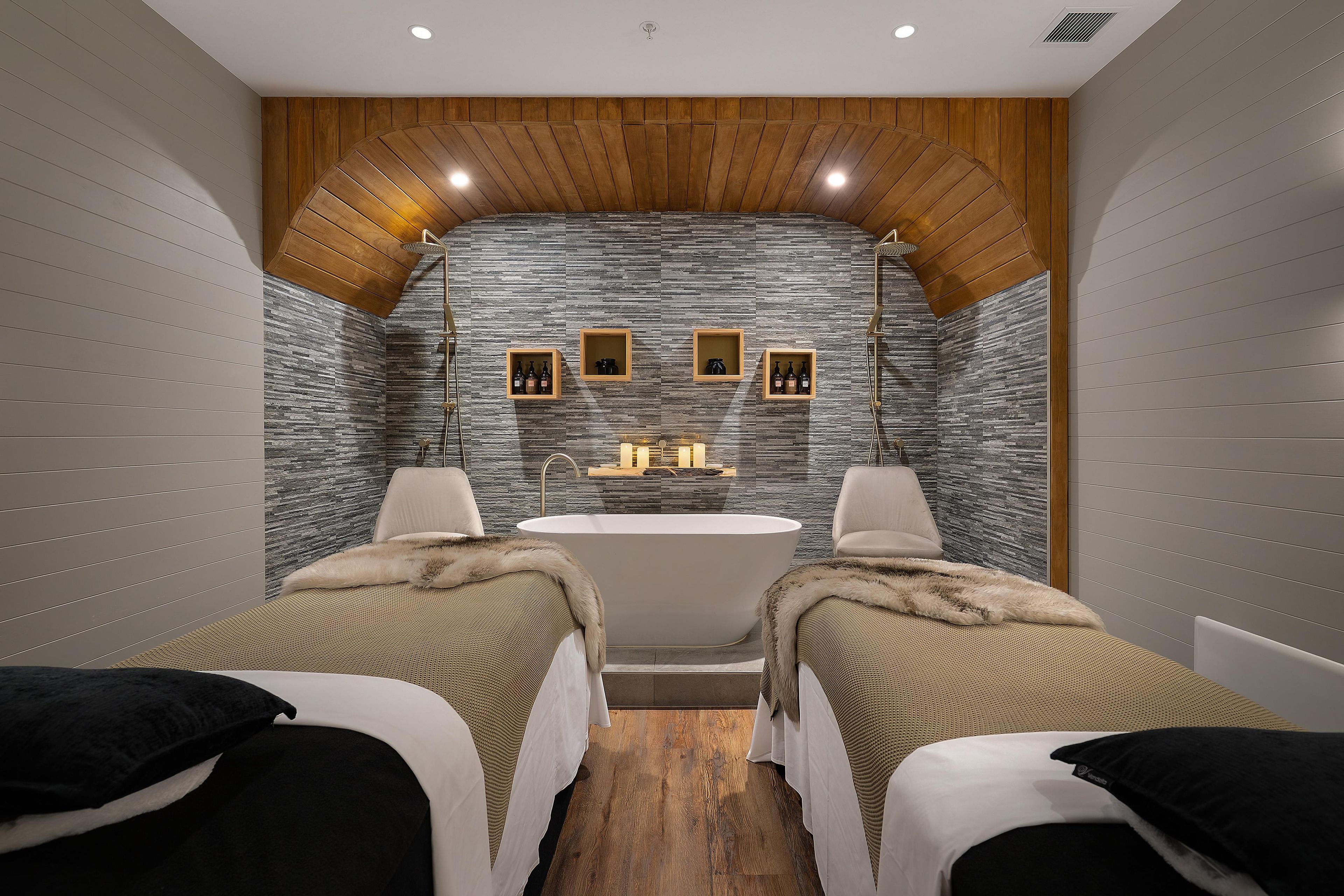 A modern bathroom with a freestanding bathtub, two beds with fur throw blankets, and wooden paneling on the ceiling.