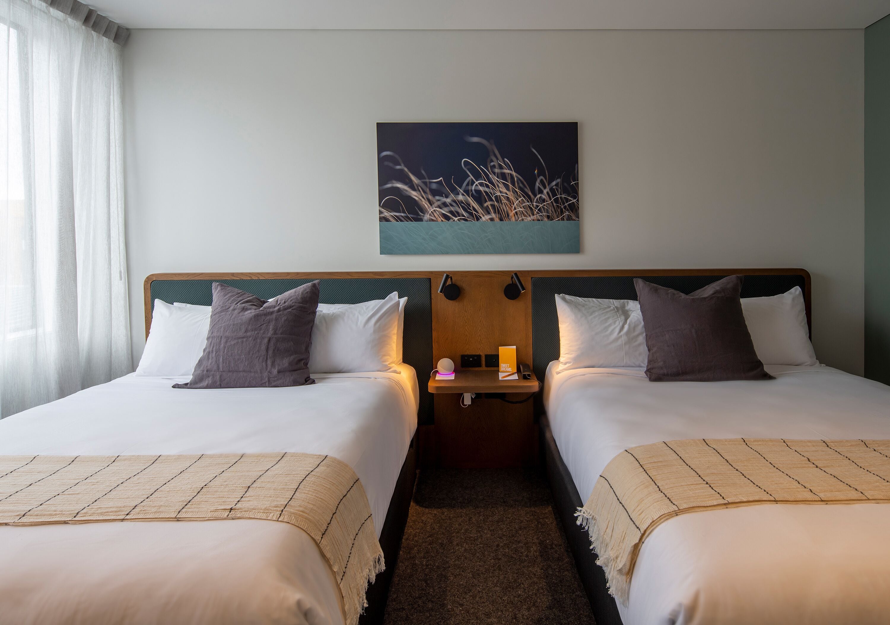 A hotel room with two beds, a nightstand, a wall-mounted artwork, and a window with curtains.