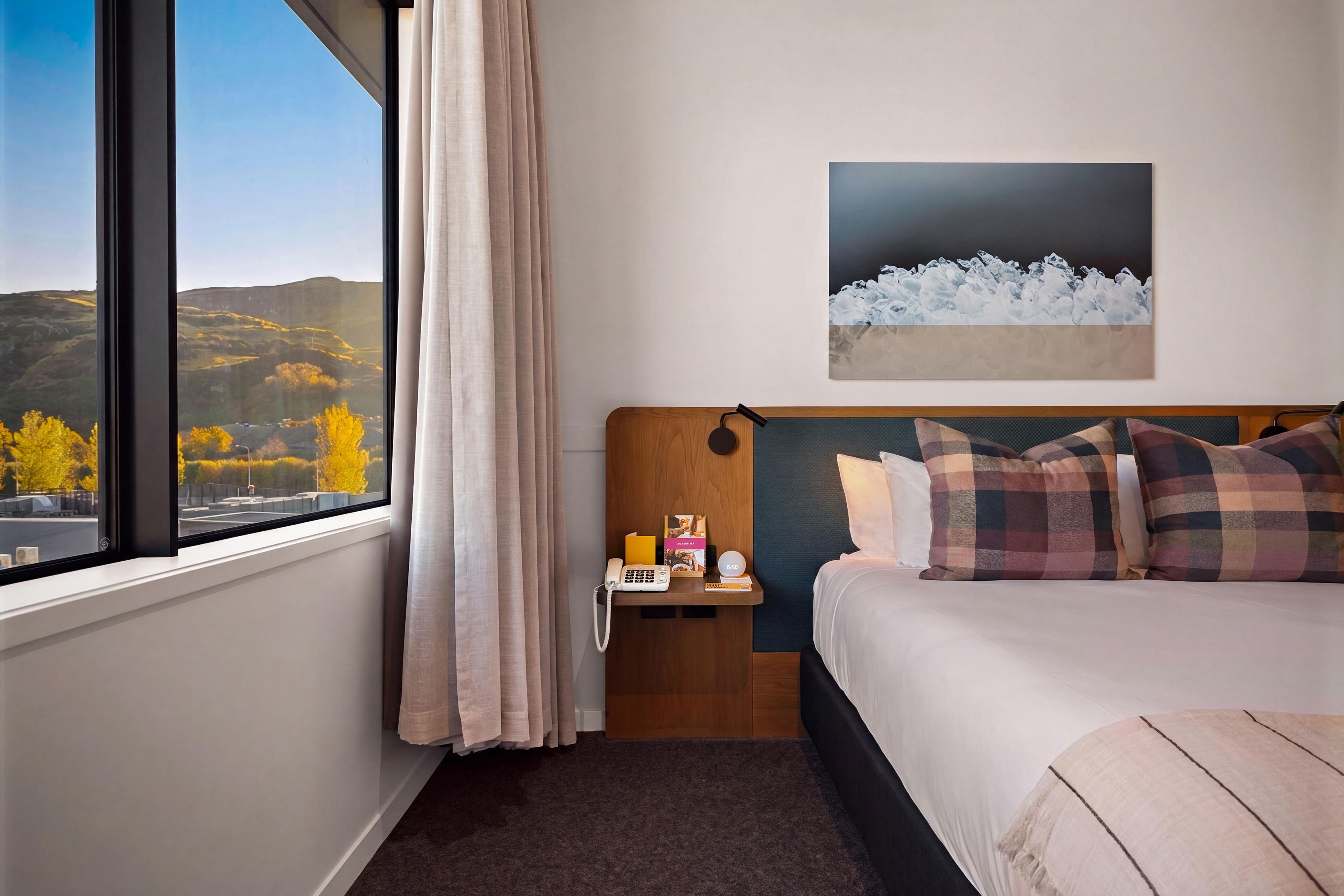 Habitación de hotel con cama, mesita de noche, un cuadro y vistas al exterior.