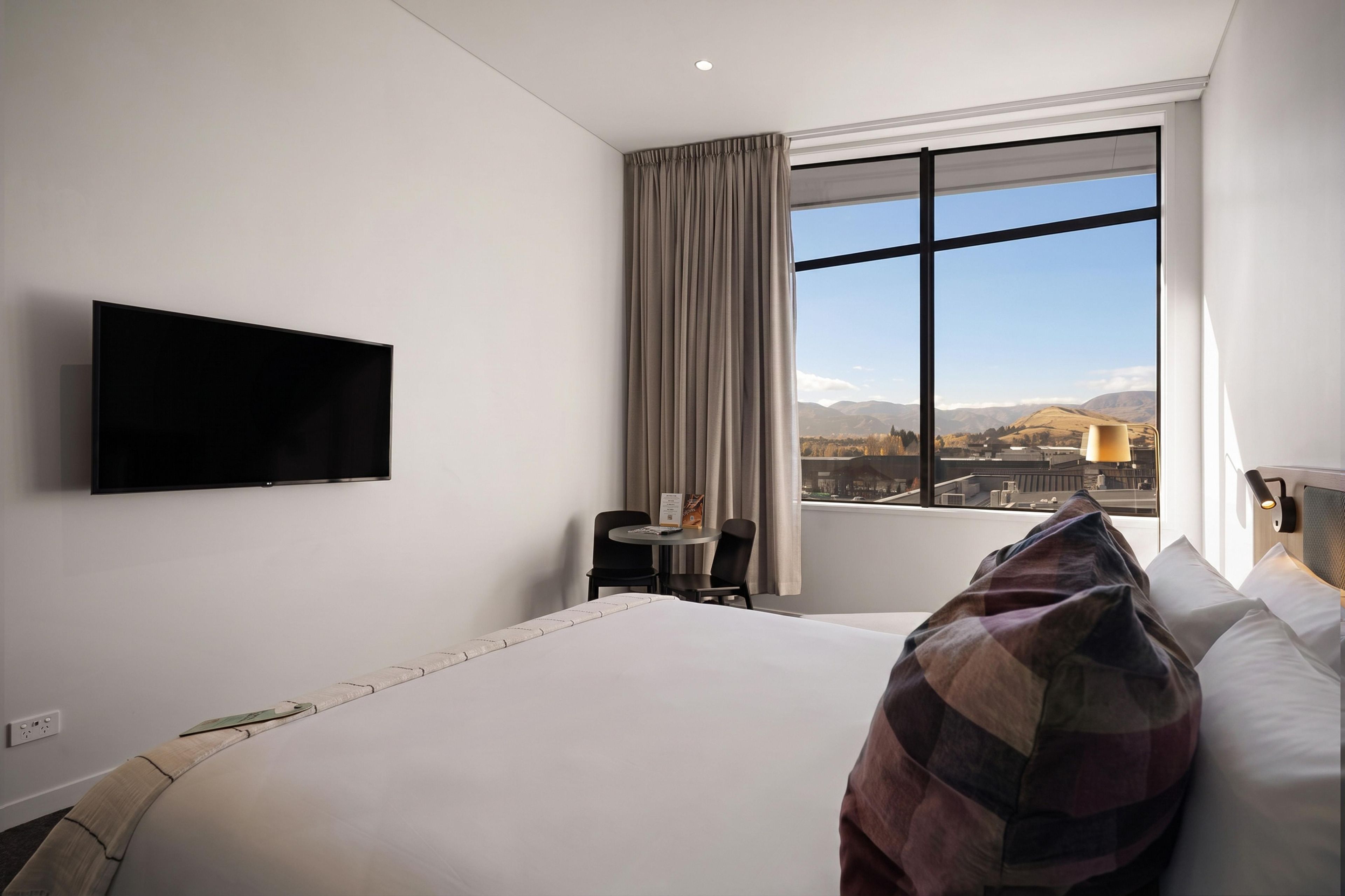A hotel room with a bed, a TV mounted on the wall, a small table with chairs, and a window with a view of the cityscape.