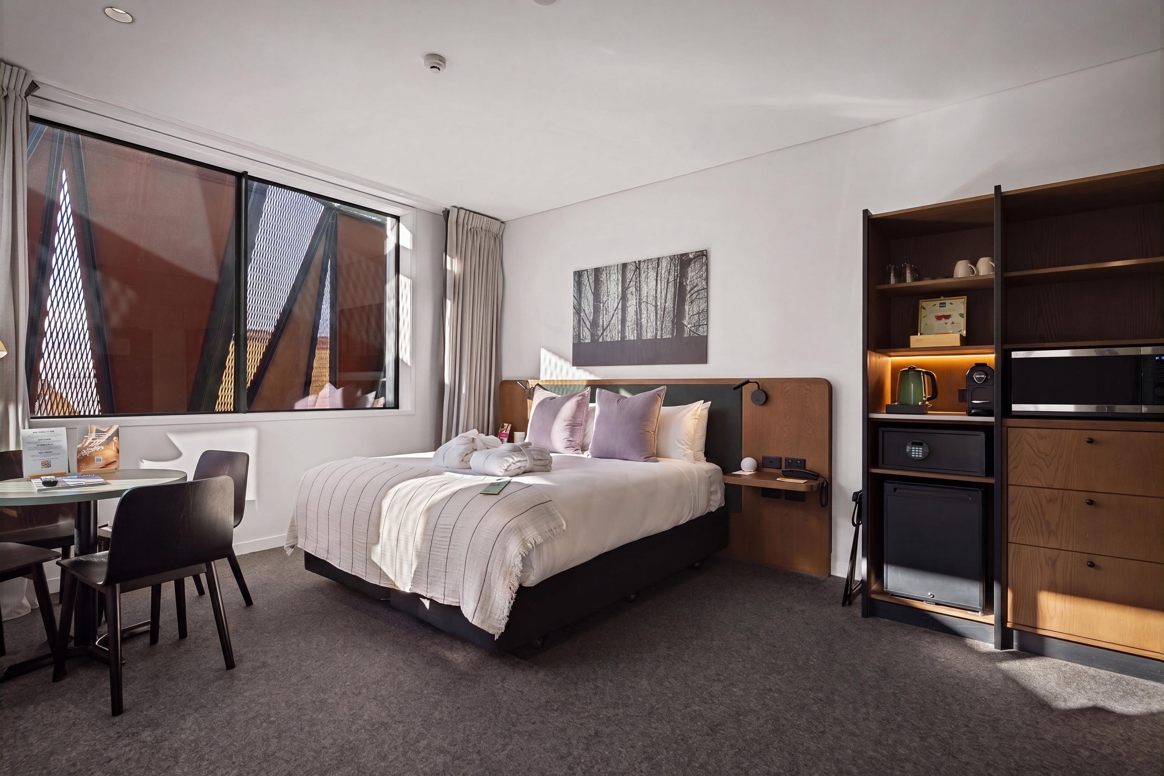 A hotel room with a bed, a desk, a chair, a bookshelf, and a painting on the wall.