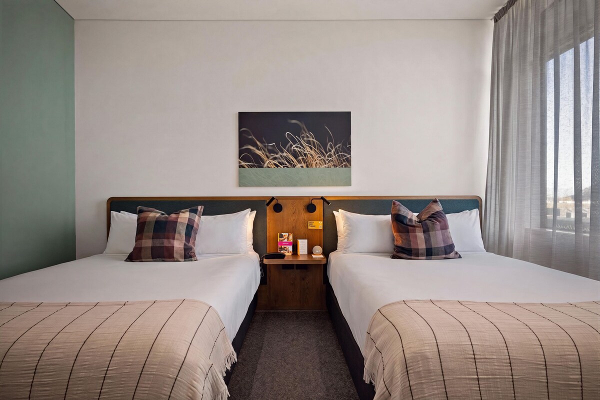A hotel room with two beds, a nightstand, a painting, and a window with curtains.