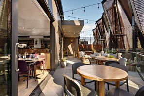 An outdoor dining area with tables and chairs, a bar with bottles and glasses, and a view of the city skyline. - Sudima Queenstown Five Mile (Queenstown)