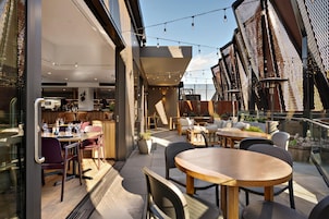 Un área de comedor al aire libre con mesas y sillas, un bar con botellas y vasos, y vistas del horizonte de la ciudad.