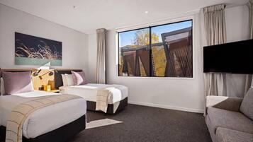 A hotel room with two beds, a sofa, a TV, and a large window with a view of trees and a fence.