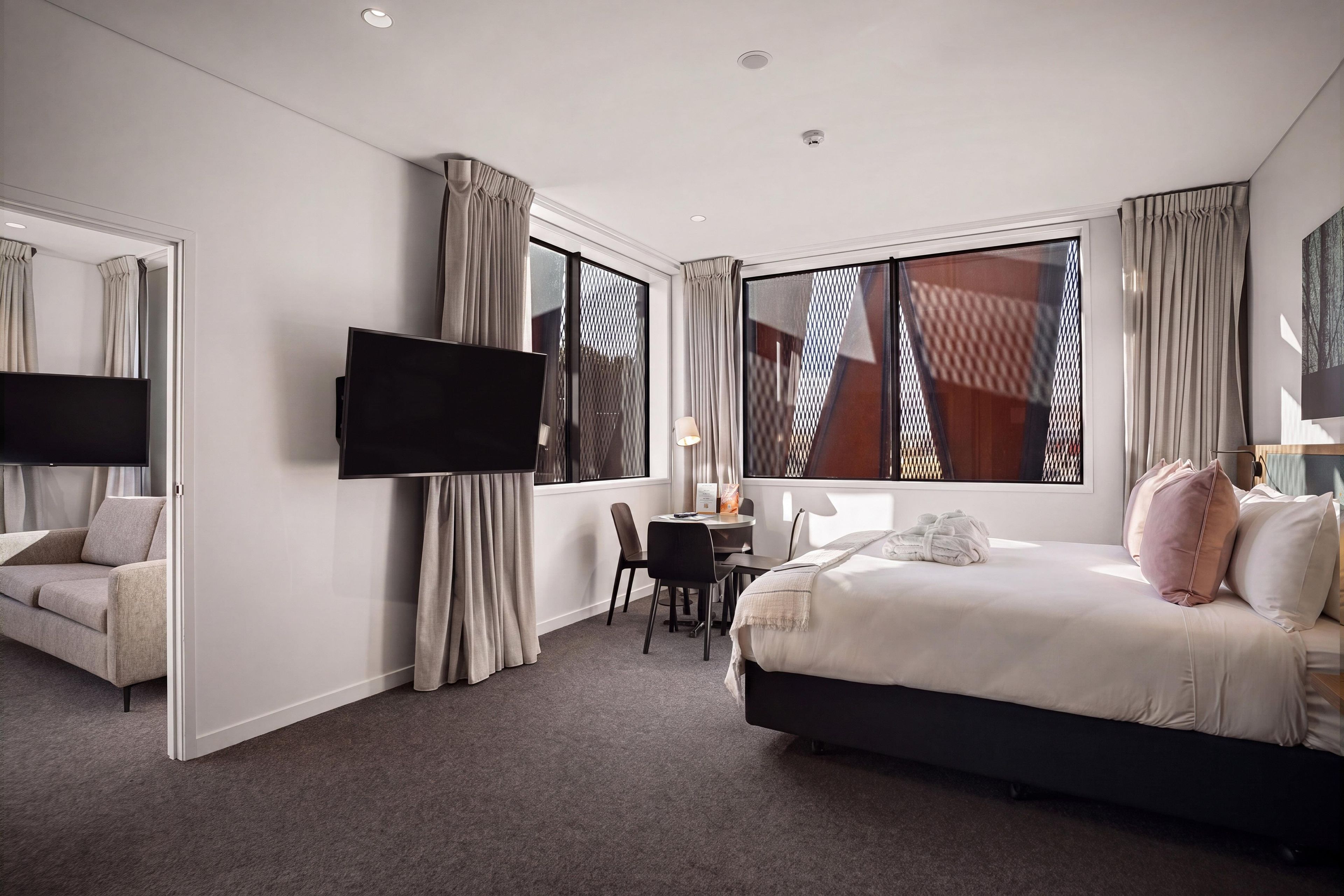 A hotel room with a bed, a desk, a chair, a TV, and large windows.