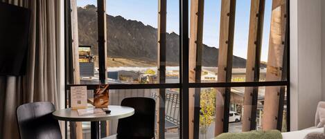 A hotel room with a large window, a bed, two chairs, a small table, and a view of mountains.