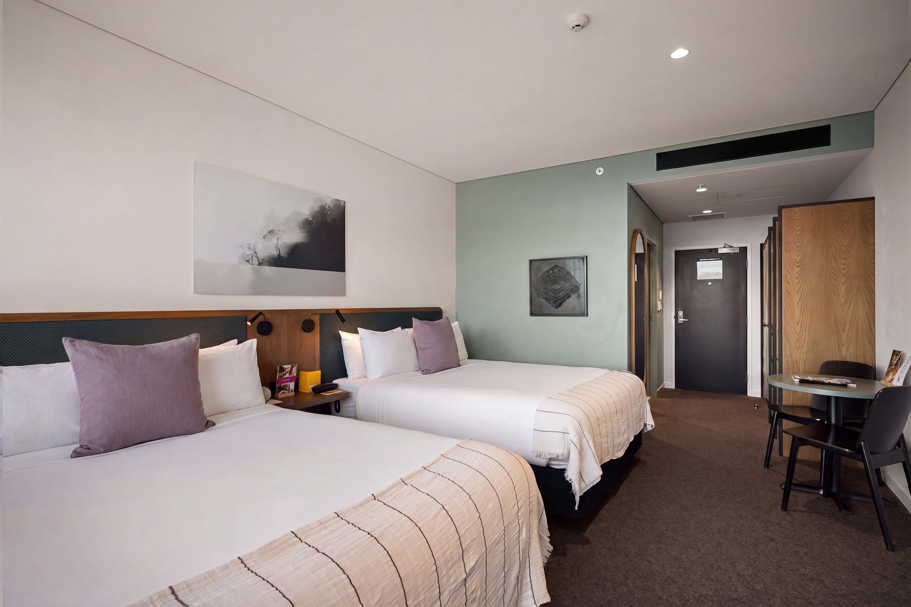 Habitació d'hotel amb dos llits, una taula de treball i una porta que dona a un passadís.