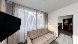 A hotel room with a sofa, a flat-screen TV, sheer curtains, a lamp, and a kitchenette. - Sudima Queenstown Five Mile (Queenstown)