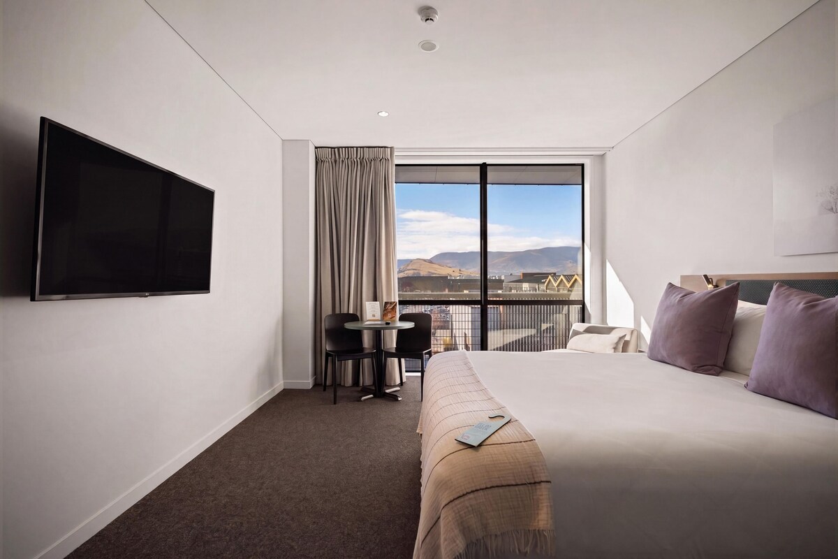 A hotel room with a large bed, a TV mounted on the wall, a small table with chairs, and a view of the mountains through a large window.