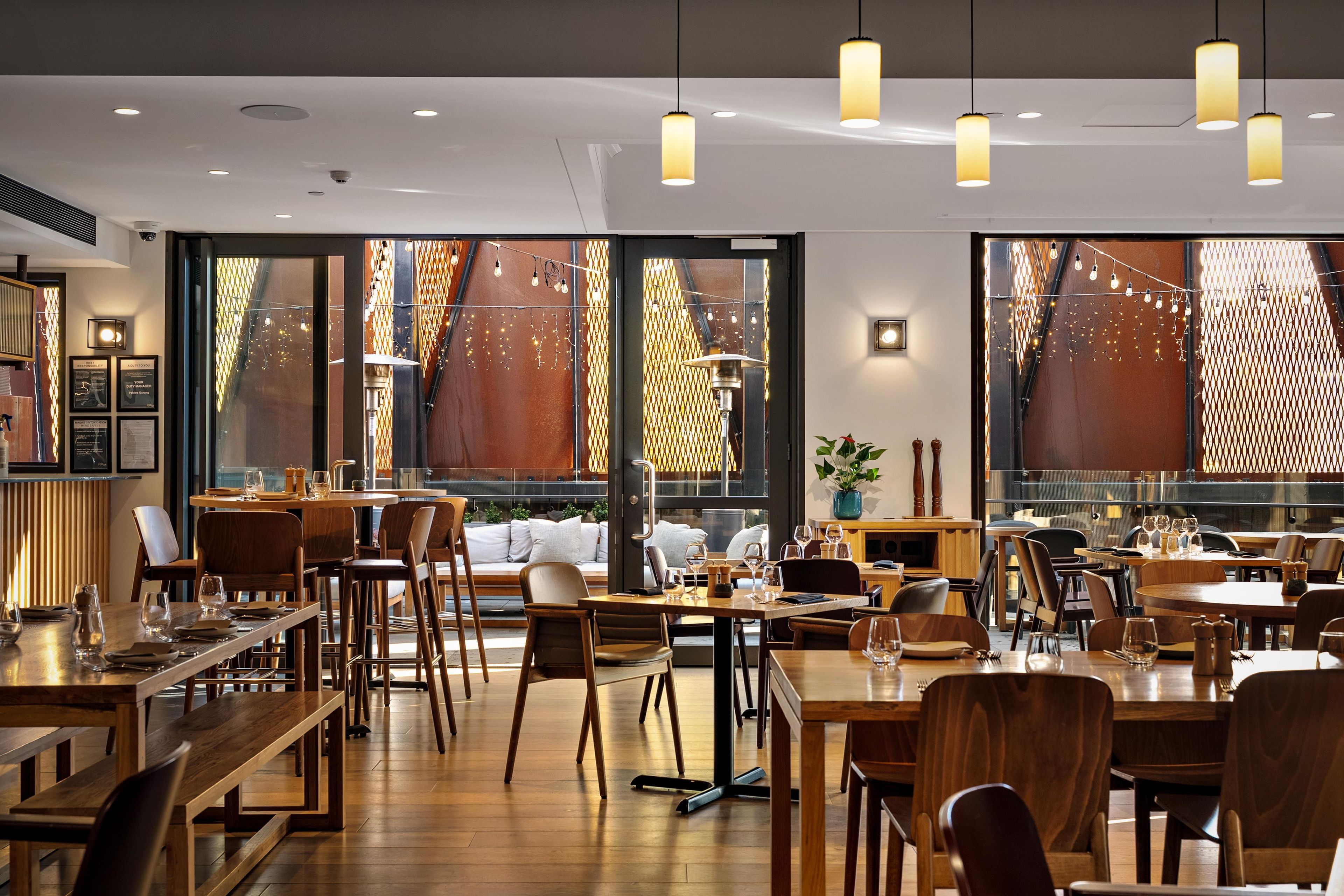 Et restaurantinteriør med flere spiseborde, stole, hængende lamper og store vinduer med dekorativt gitter.