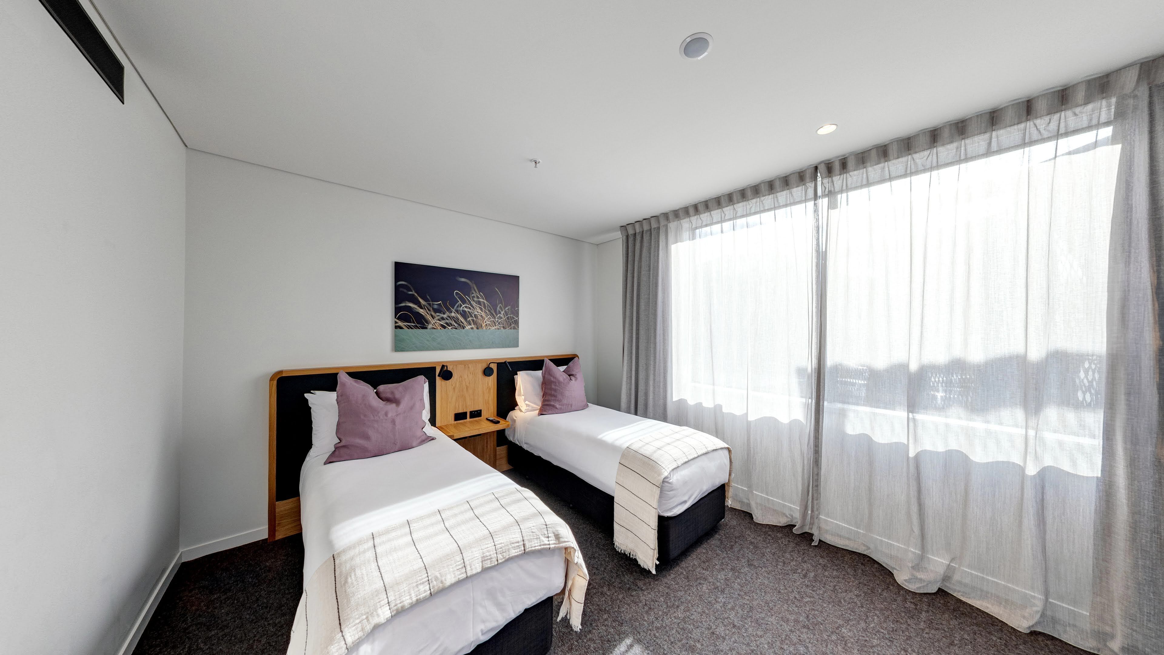 A hotel room with two single beds, a large window with sheer curtains, a ceiling light, and a painting on the wall.