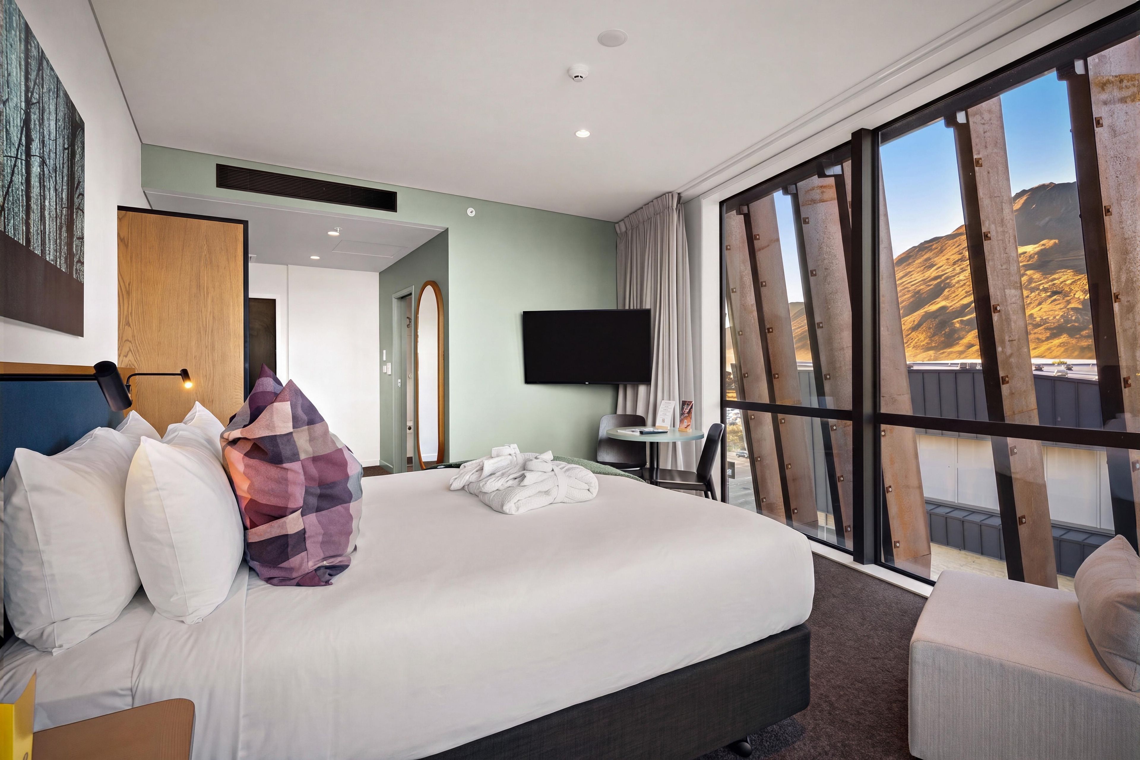 A hotel room with a large bed, a desk, a chair, a TV, and a view of a mountainous landscape.