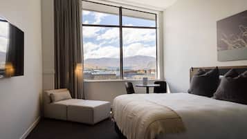 Habitación de hotel con cama, sofá, mesa y silla. Se ve un ventanal con vistas a montañas y al paisaje urbano.