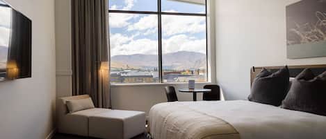 A hotel room with a bed, a sofa, a table, and a chair. There is a window with a view of mountains and a cityscape.