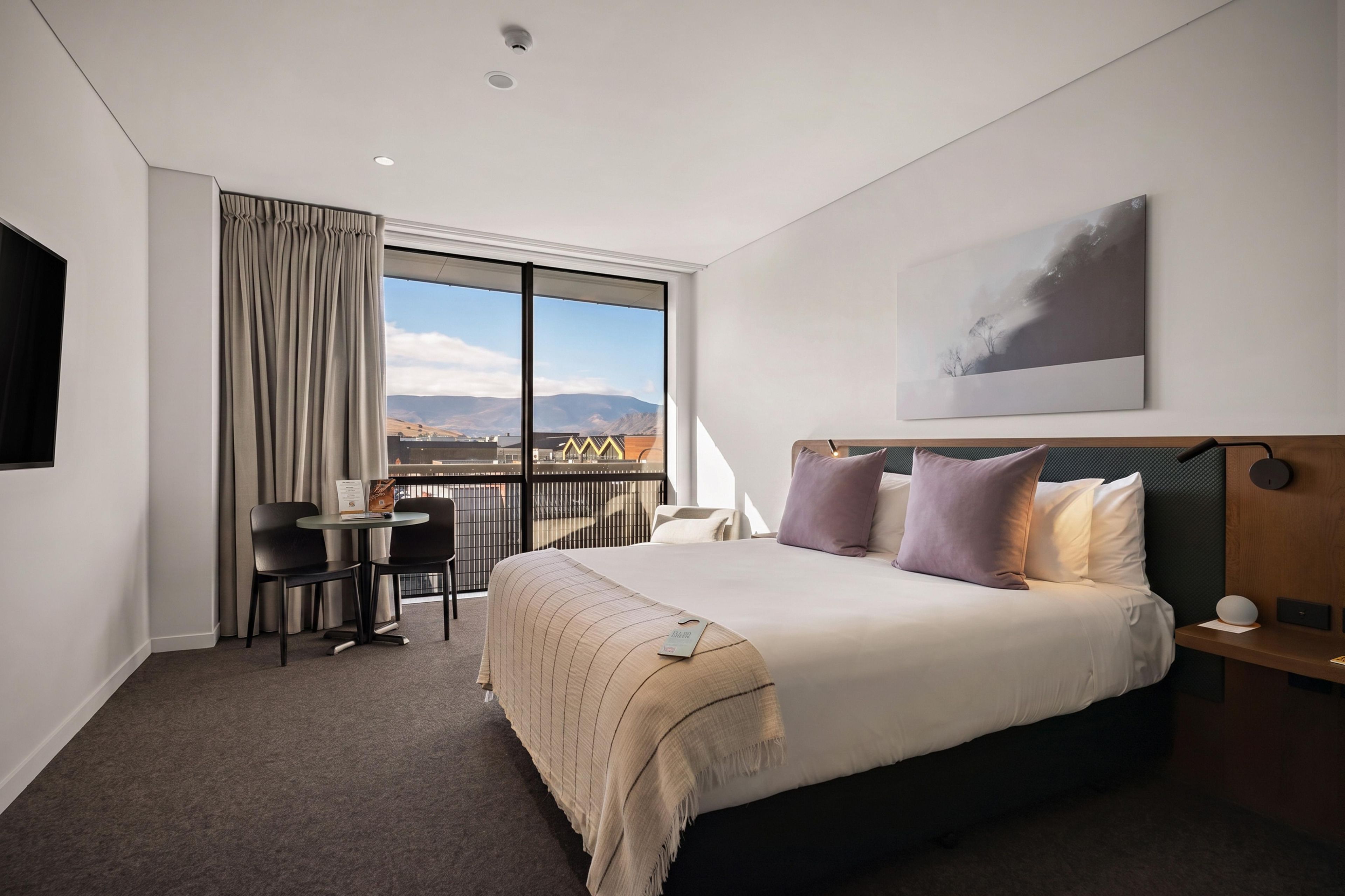 A hotel room with a large bed, a desk with chairs, a balcony with a view, and a mounted artwork.