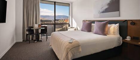 Habitación de hotel con una cama grande, un escritorio con sillas, un balcón con vistas y una obra de arte colgada en la pared.
