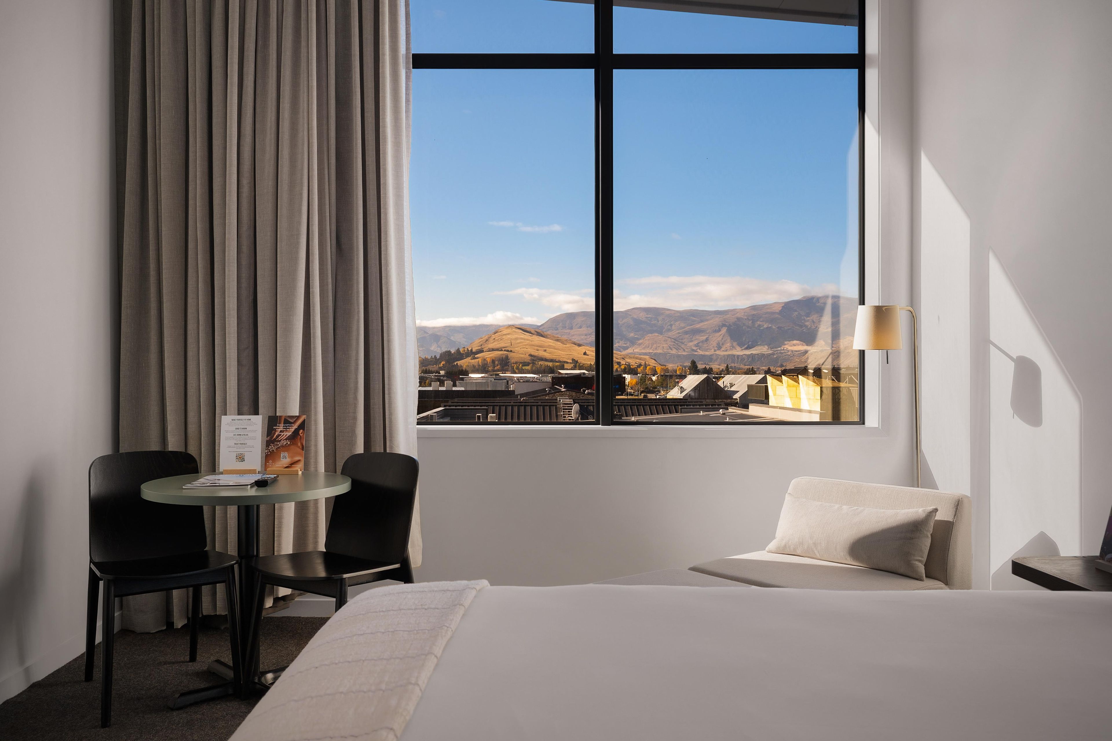 Habitación de hotel con cama, escritorio con sillas, ventana y vistas a la montaña, además de una lámpara.