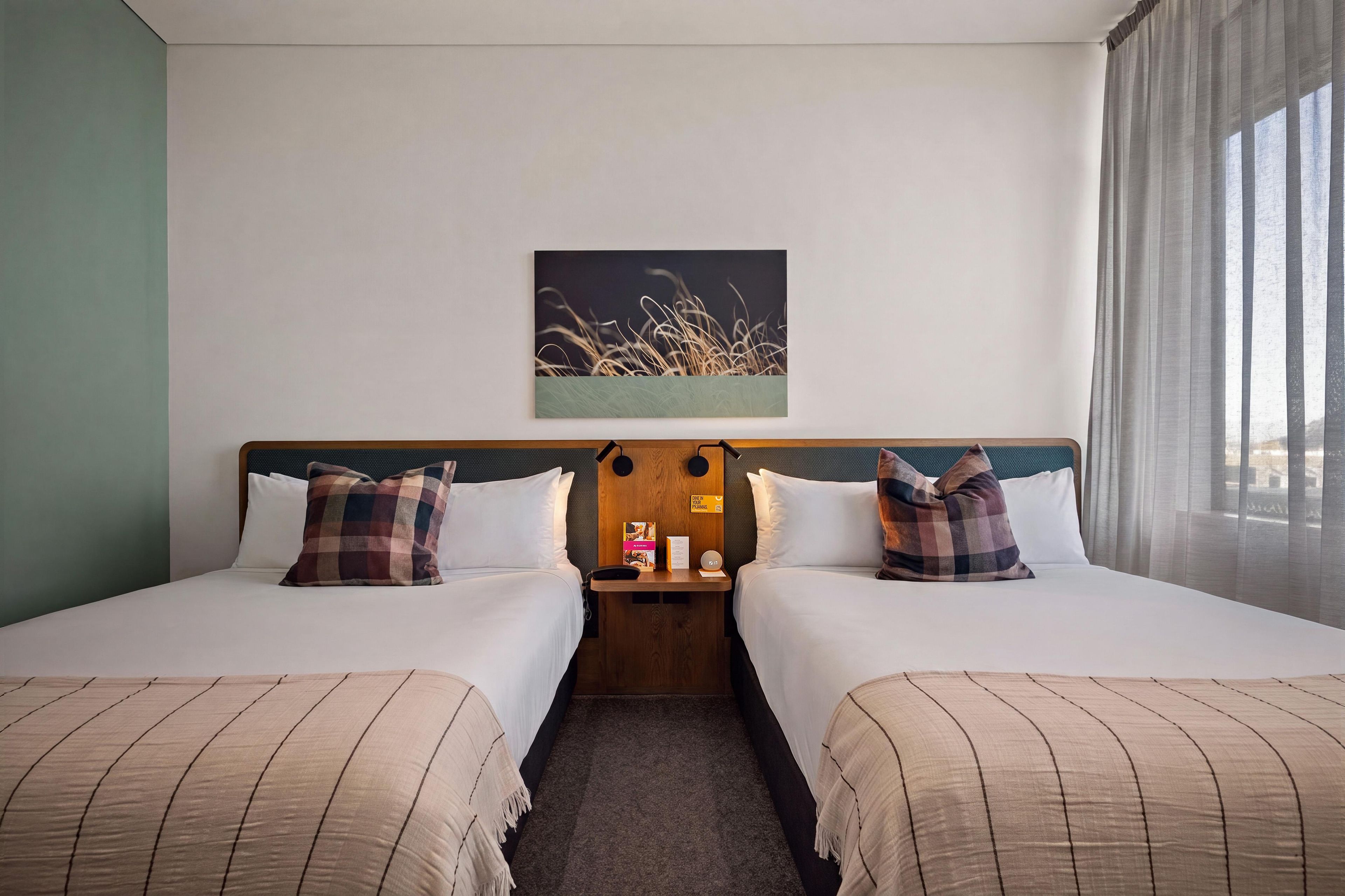 Une chambre d’hôtel avec deux lits, une table de chevet, un tableau et une fenêtre avec des rideaux.