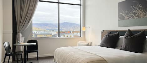 A hotel room with a large window, a bed, a desk with a chair, and a painting on the wall.
