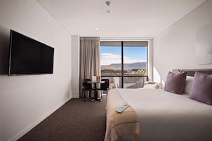 A hotel room with a large bed, a TV mounted on the wall, a small table with chairs, and a view of the mountains through a large window.