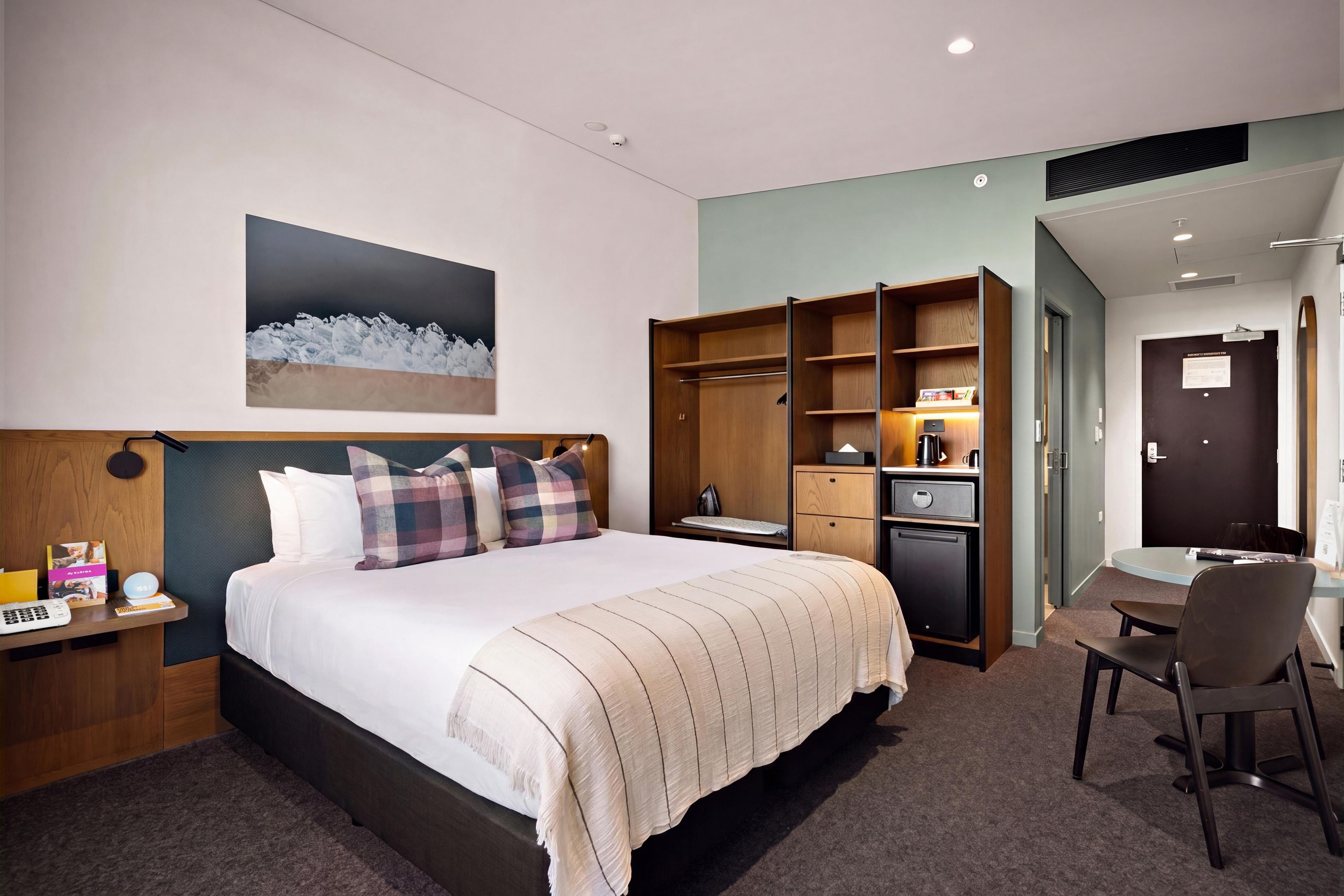A hotel room with a bed, a desk, a chair, a wardrobe, and a painting on the wall.