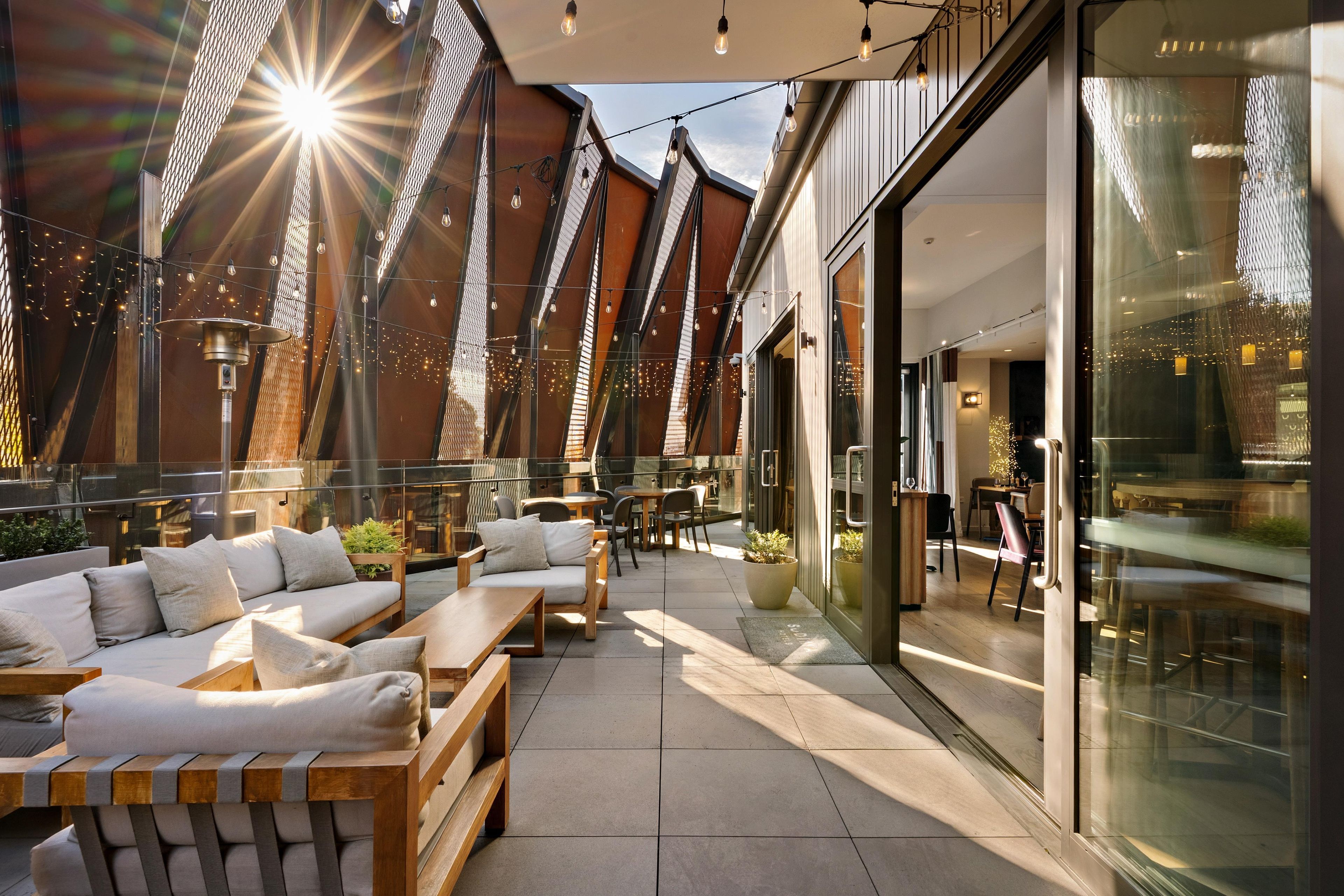 Un espacio al aire libre con bancos de madera y cojines, un muro de vidrio y guirnaldas de luces.