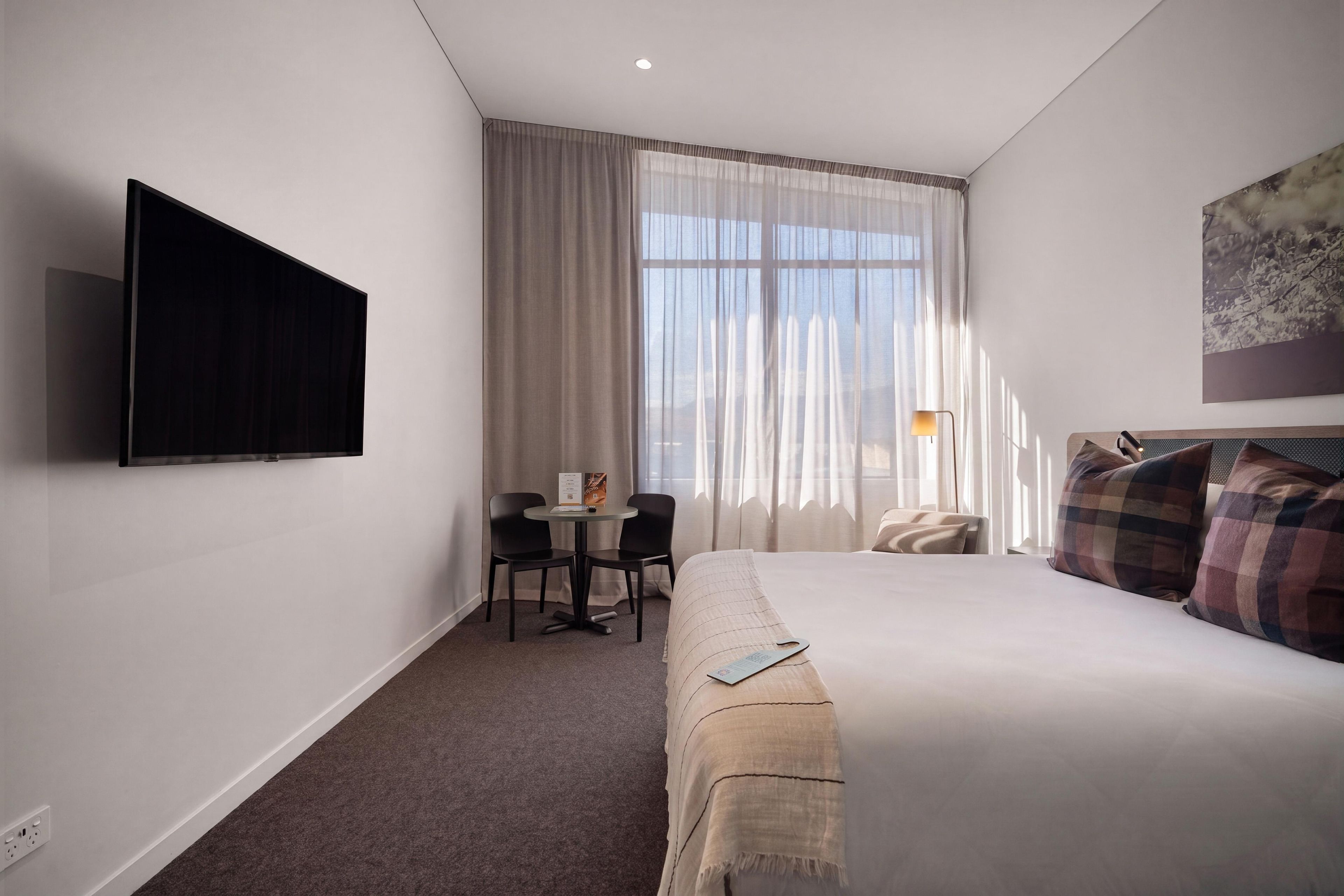A hotel room with a bed, a desk with chairs, a flat-screen TV, a window with curtains, and a painting on the wall.