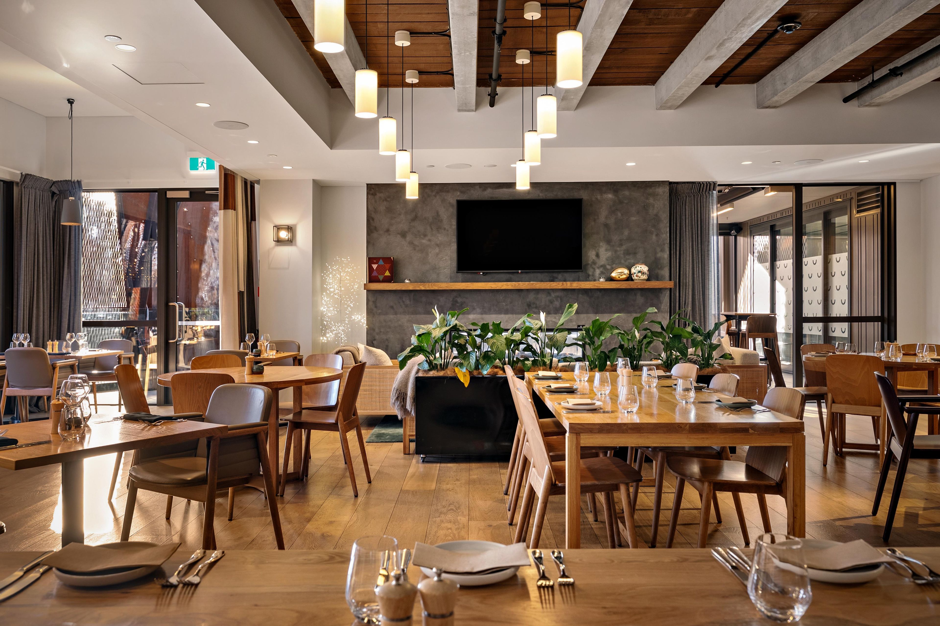 A modern dining area with wooden tables and chairs, a flat-screen TV mounted on the wall, and potted plants.