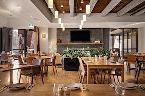 A modern dining area with wooden tables and chairs, a flat-screen TV mounted on the wall, and potted plants. - Sudima Queenstown Five Mile (Queenstown)