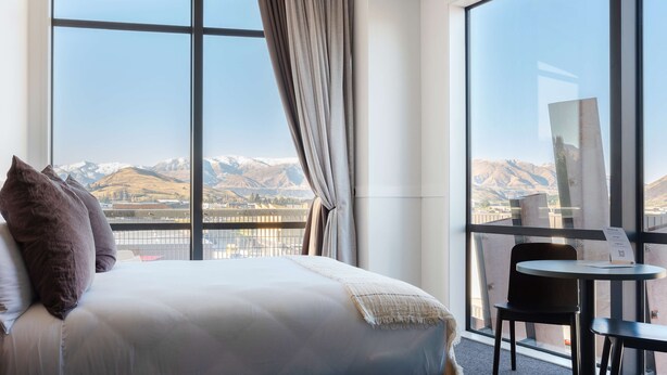 A hotel room with a large bed, a small table, and a chair. There is a large window with a view of mountains. - Sudima Queenstown Five Mile (Queenstown)
