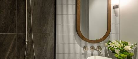 A bathroom with a shower, a sink, a mirror, and a vase of flowers.