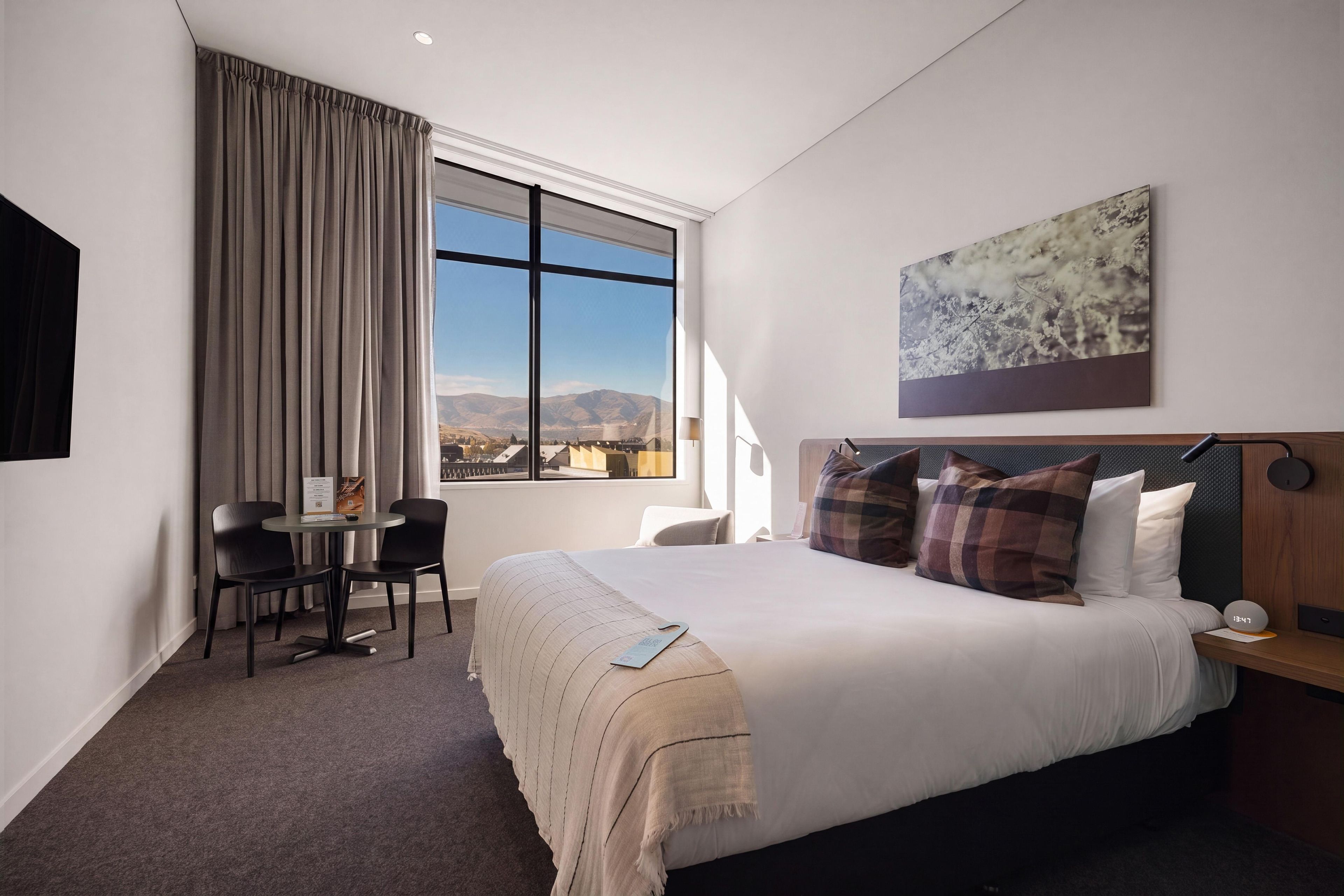 Una habitación de hotel con una cama, un escritorio, dos sillas, un ventanal con vista a las montañas y un cuadro en la pared.
