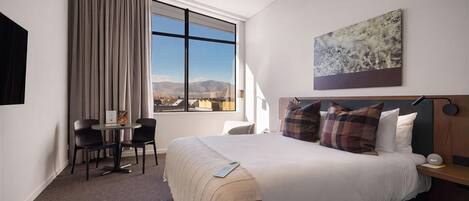 Una habitación de hotel con una cama, un escritorio, dos sillas, un ventanal con vista a las montañas y un cuadro en la pared.