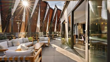 Un espacio al aire libre con bancos de madera y cojines, un muro de vidrio y guirnaldas de luces.