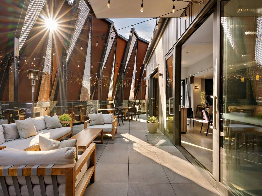 Un espacio al aire libre con bancos de madera y cojines, un muro de vidrio y guirnaldas de luces.