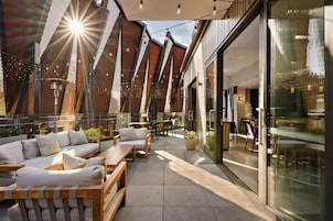 Un espacio al aire libre con bancos de madera y cojines, un muro de vidrio y guirnaldas de luces.