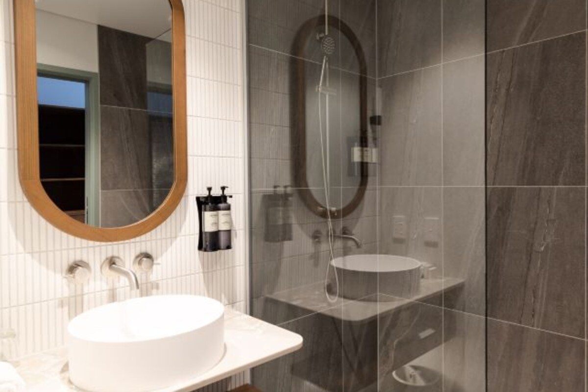 A modern bathroom with a round mirror, a white sink, a shower, and a marble-tiled wall.