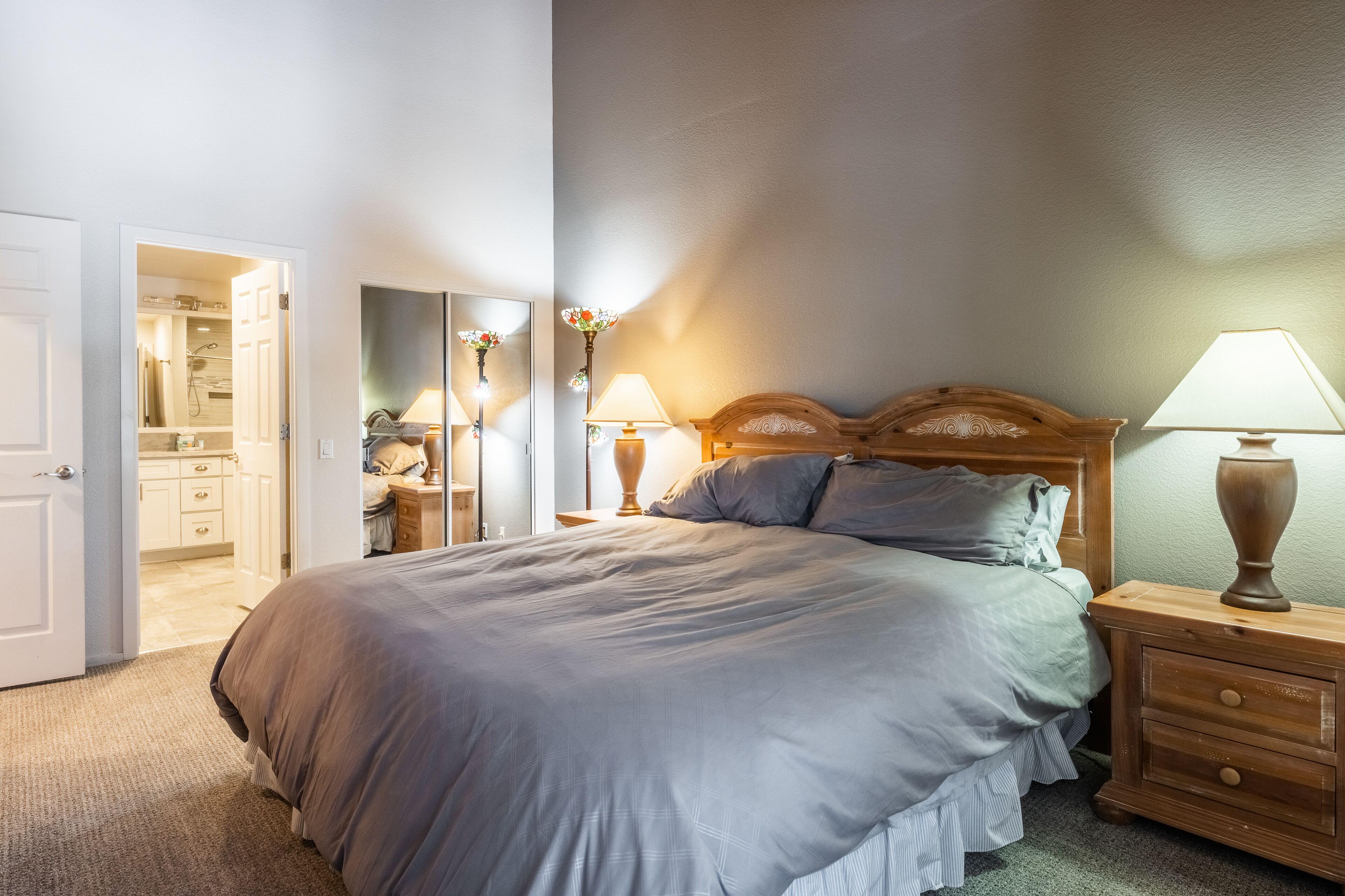 A bedroom with a large bed, wooden headboard, two bedside tables with lamps, a mirror, and an open door leading to a bathroom.