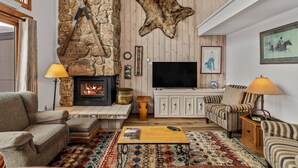 Ein gemütliches Wohnzimmer mit Kamin aus Naturstein, einem großen Fernseher und rustikalen Einrichtungsgegenständen, darunter ein aufgehängtes Tierfell.