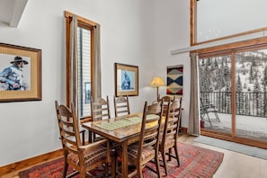 A dining area with a wooden table and chairs, framed artwork on the walls, and a view of a snowy landscape through a large sliding glass door. - Ski In/Out-Pool-HotTub-Views-Grill-Just Remodeled (Steamboat Springs)