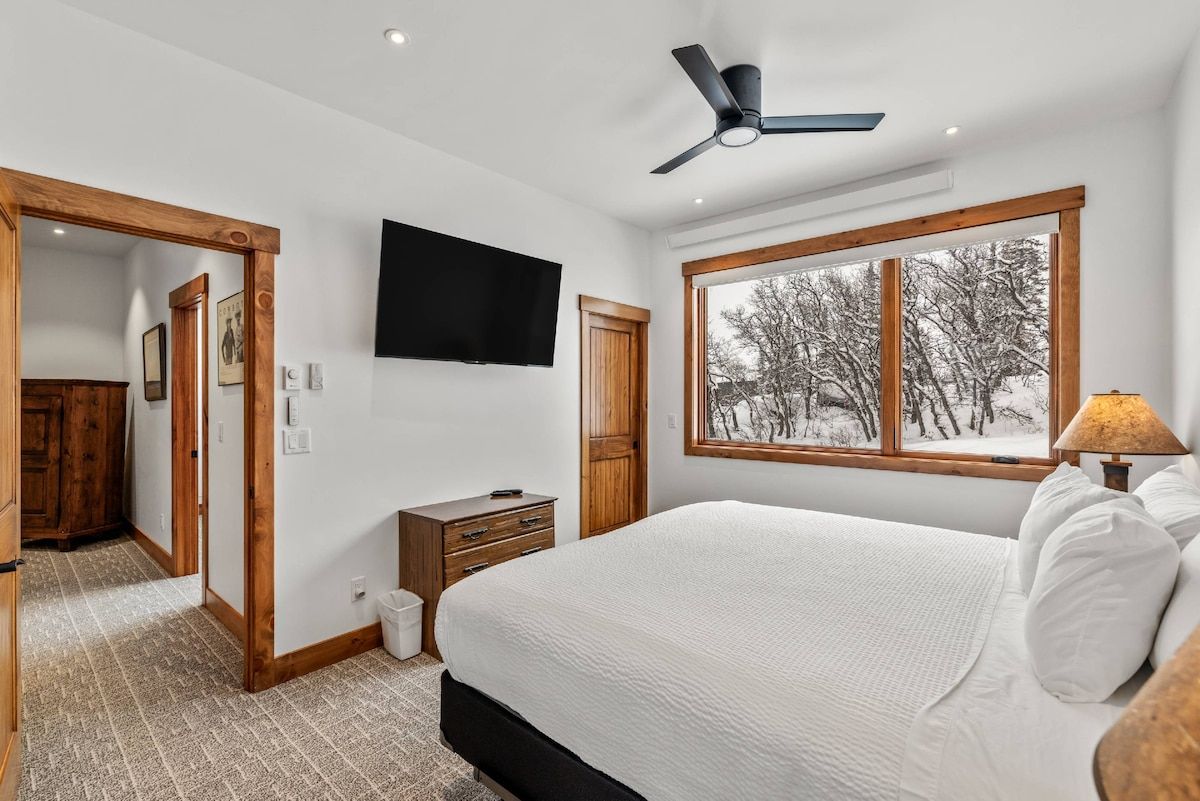 A bedroom with a bed, a ceiling fan, a TV, a window with a view of trees, and a wooden door.