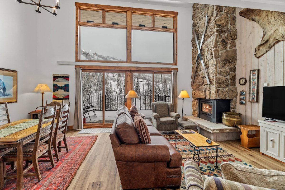 A cozy living room with a fireplace, a dining area with a wooden table and chairs, and a large window with a view of the outdoors.