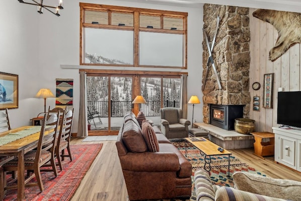 A cozy living room with a fireplace, a dining area with a wooden table and chairs, and a large window with a view of the outdoors.