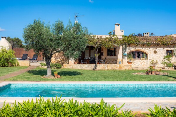 A poolside view of a house with a stone facade, outdoor seating, and a clear blue sky. - Holiday Home "Ca Sa Padrineta" with Wi-Fi, Air Conditioning, Pool, Garden & Terrace (Capdepera)