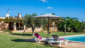 Ein Swimmingpool mit Liegestühlen und einem Sonnenschirm, umgeben von einem gepflegten Rasen und einem Steinhaus mit Satellitenschüssel.