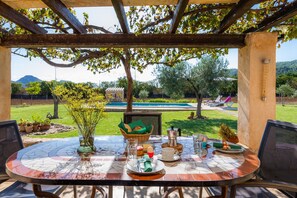 An outdoor patio with a round table set for dining, surrounded by chairs, with a view of a pool and garden. - Holiday Home "Ca Sa Padrineta" with Wi-Fi, Air Conditioning, Pool, Garden & Terrace (Capdepera)